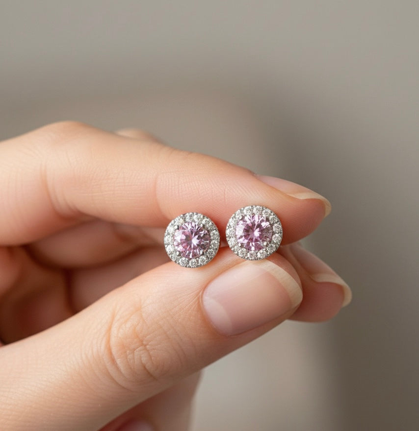 Detailansicht von zwei rhodinierten Halo-Ohrsteckern aus 925er Sterling Silber mit rosa Zirkonia-Stein, präsentiert zwischen den Fingern einer Hand.