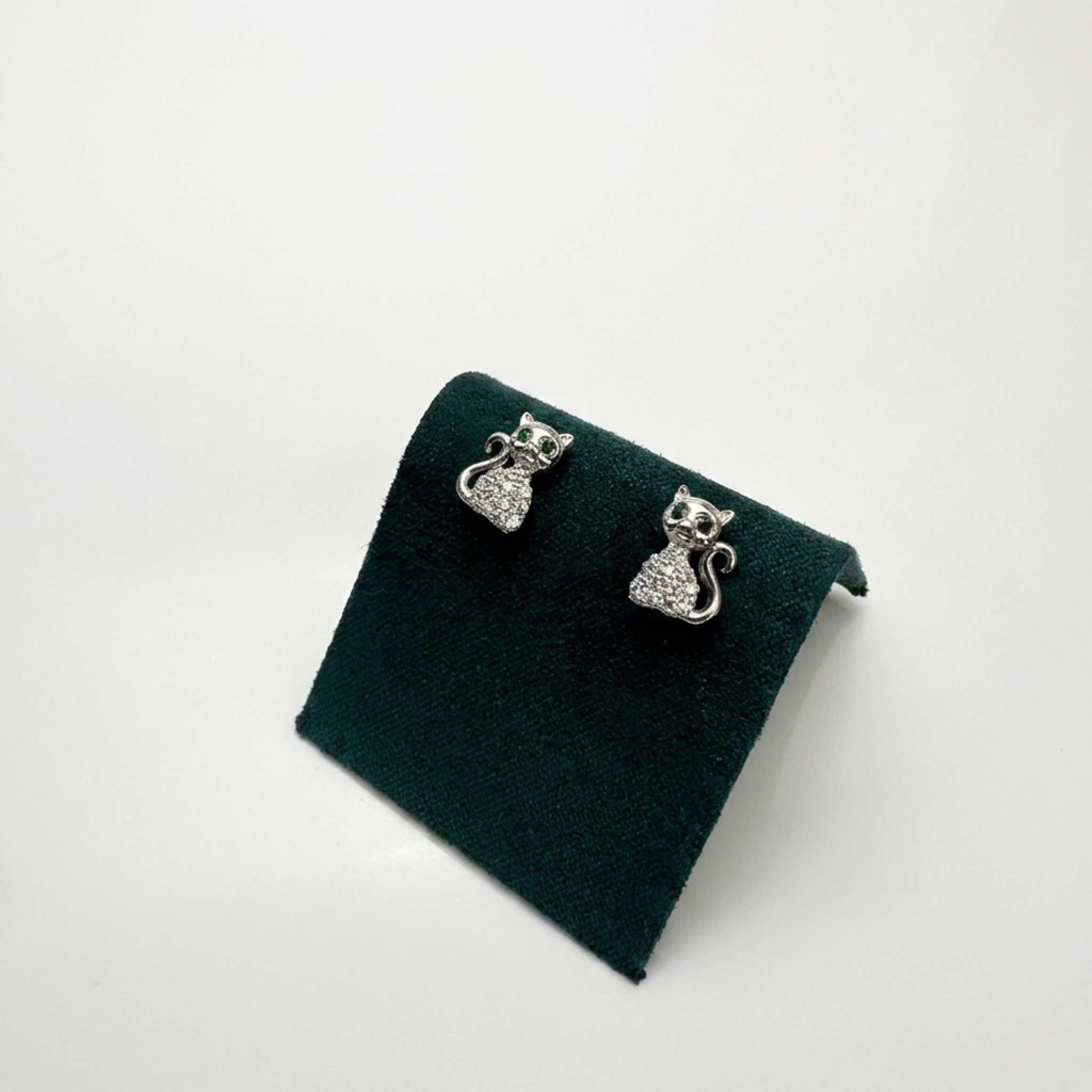 Handgefertigter Designerschmuck: Rhodinierte Motiv-Ohrringe "Katze" aus 925 Sterling Silber mit klaren und grünen Zirkonia auf einer exklusiven dunkelgrünen Samtpräsentation.
