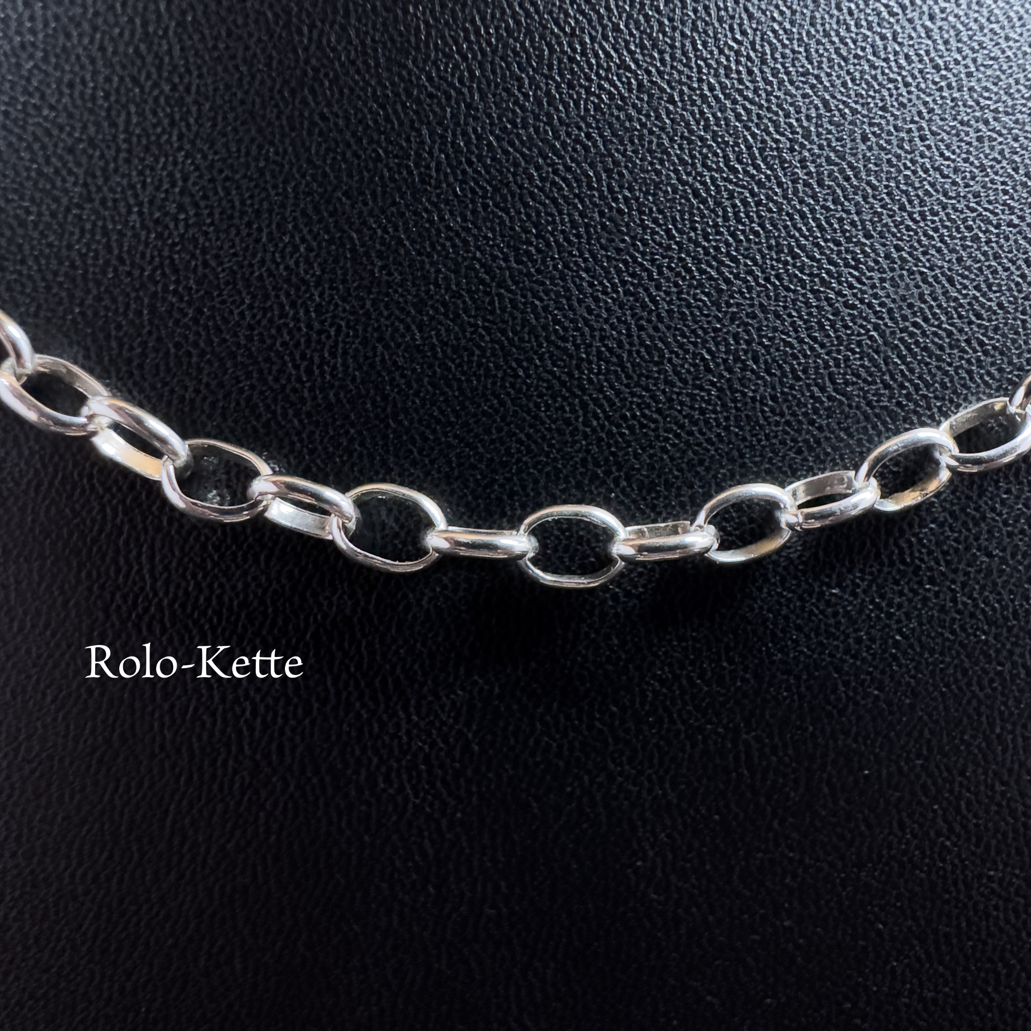 Nahaufnahme einer filigranen Rolo-Kette aus glänzendem 925er Silber auf schwarzem Lederhintergrund mit dem Schriftzug Rolo-Kette.