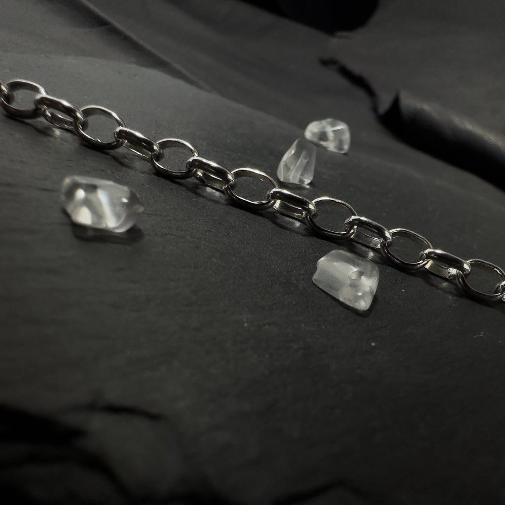 Detailaufnahme einer silbernen Gliederkette im Rolo-Stil, die sich von einem runden Endring im Vordergrund über eine strukturierte, dunkle Steinfläche nach hinten erstreckt.