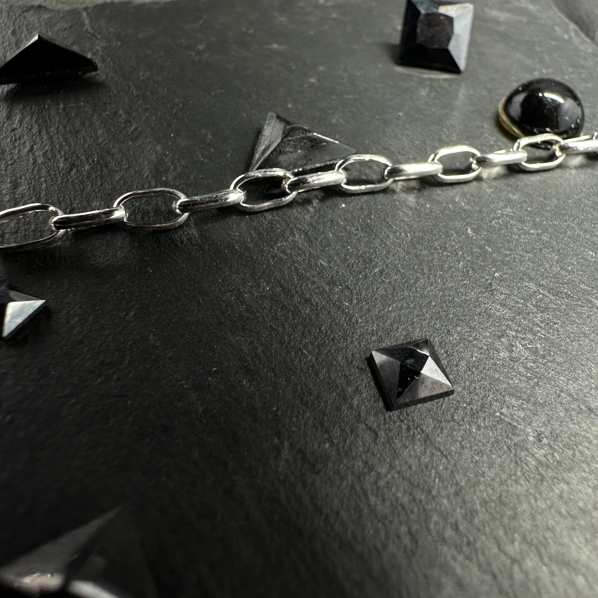 Makroaufnahme einer Rolo-Kette (Ankerkette) aus 925er Silber auf einer dunklen Steinoberfläche, dekoriert mit kleinen, pyramidenförmigen schwarzen Schmuckelementen.